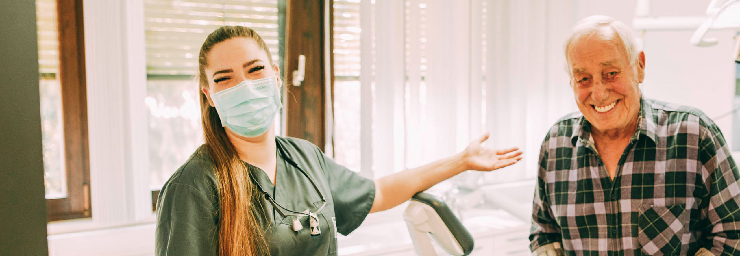 Zahnärztin Ricarda Richter lacht hinter dem Mundschutz und zeigt auf einen lachenden Patienten mit perfekten Zähnen.