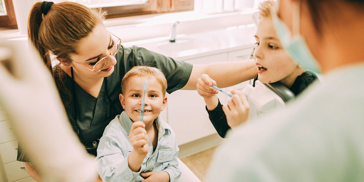 Gruppenbild mit Kindern: Zahnärztin Ricarda Richter spricht im Gespräch mit zwei Jungs, etwa 5 und acht Jahre alt. Sie erklärt das richtige Zähneputzen.